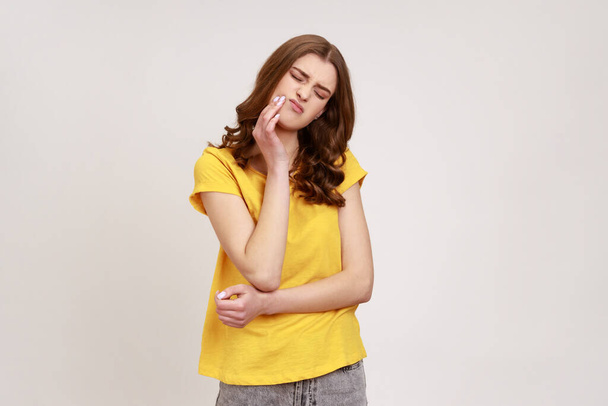 Frustrated wavy haired teenager girl wearing yellow casual T-shirt touching cheek and wincing in pain feeling terrible tooth ache, periodontal disease. Indoor studio shot isolated on gray background. - Fotografie, Obrázek