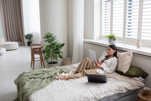 Νεαρή με αυτοπεποίθηση Ασιάτισσα με casual ρούχα που κάθεται στο κρεβάτι και διαβάζει εφημερίδες - Φωτογραφία, εικόνα