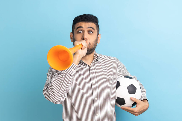 Extremely positive satisfied businessman with bullhorn holding soccer ball, celebrating victory of favorite football team, wearing striped shirt. Indoor studio shot isolated on blue background. - Photo, Image