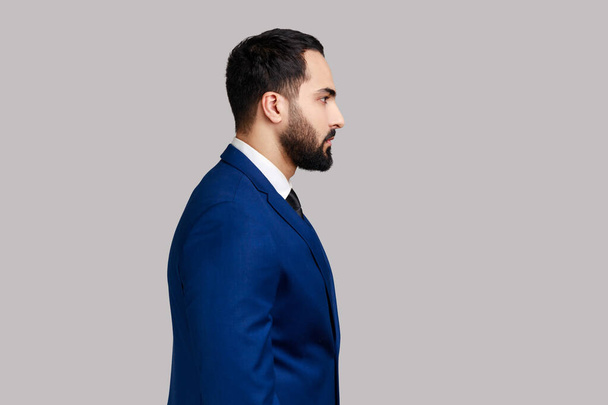 Side view portrait of confident bearded male looking at camera with serious expression, unsmiling determined business man, wearing official style suit. Indoor studio shot isolated on gray background. - Zdjęcie, obraz
