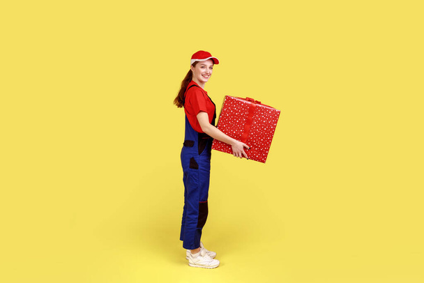 Side view portrait of courier woman standing with big present box in hands, looking smiling at camera, fast delivery door-to-door, wearing overalls. Indoor studio shot isolated on yellow background. - Zdjęcie, obraz