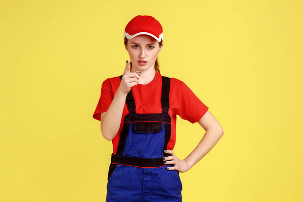 Portrait of bossy confident worker woman standing, raising finger, showing warning gesture, looking at camera, wearing overalls and red cap. Indoor studio shot isolated on yellow background. - Фото, изображение