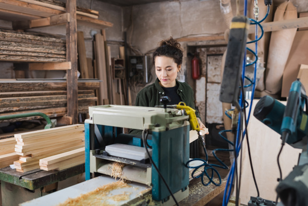 Joven carpintero de pie cerca del banco más grueso y tablones de madera en el taller  - Foto, imagen