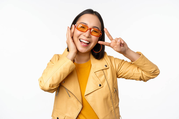 Porträt eines stilvollen asiatischen modernen Mädchens mit Sonnenbrille und gelbem Sakko, das Frieden zeigt, V-Zeichen-Geste, auf weißem Hintergrund stehend, glücklich lächelndes Gesicht - Foto, Bild