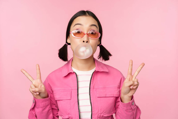 Beautiful korean girl in sunglasses, blowing bubblegum bubble and showing peace signs, standing over pink background - Foto, Imagen