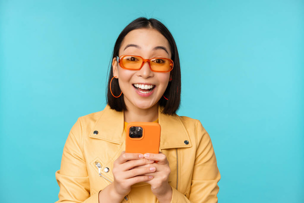 Porträt einer begeisterten asiatischen Frau mit Sonnenbrille, mit Handy, lächelnd und lachend, glücklich aussehend, Smartphone in der Hand, vor blauem Hintergrund stehend - Foto, Bild