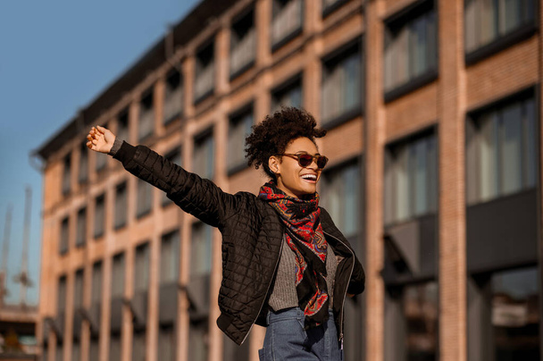 Una chica de piel oscura en gafas de sol caminando por la calle y sintiéndose alegre - Foto, imagen