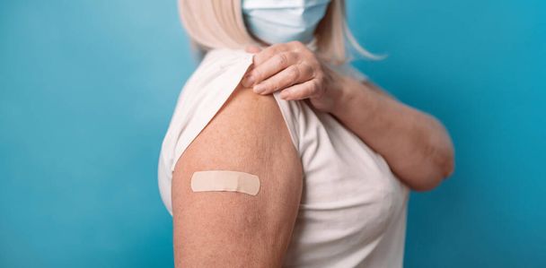 Vacunación de ancianos. Mujer mayor madura sana de 50 años mostrando brazo con yeso después de la vacuna, usando mascarilla sobre fondo azul. - Foto, imagen