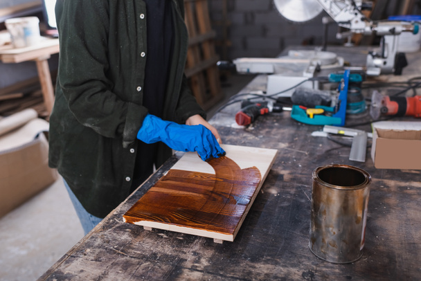 Cropped view of furniture designer in rubber glove applying wood stain on board in workshop  - Foto, afbeelding