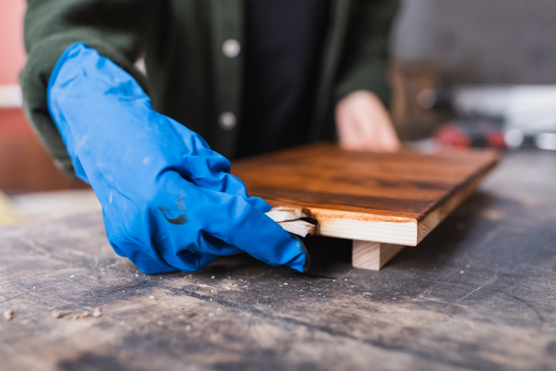 Vista recortada del carpintero en guante de goma aplicando mancha de madera a bordo  - Foto, Imagen