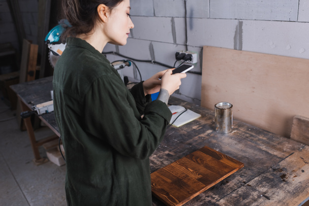 Vista lateral de la joven artesana usando smartphone cerca de tablón de madera en taller  - Foto, Imagen