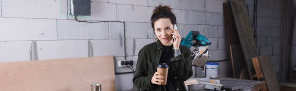 Charpentier positif tenant une tasse en papier et parlant sur smartphone en atelier, bannière  - Photo, image