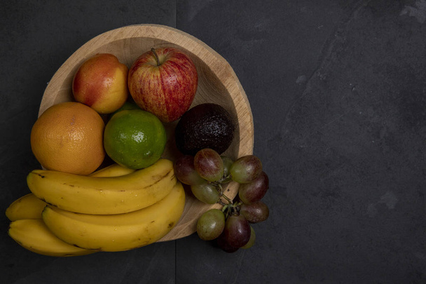 Wooden trough with assorted fruits, on black stone background. Space for text - 写真・画像