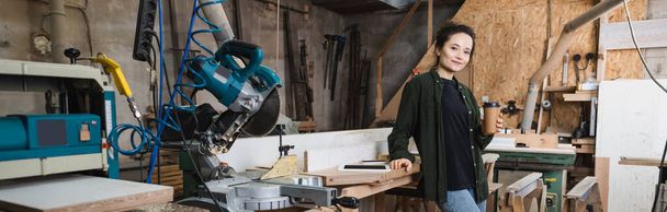 Alegre carpintero sosteniendo una taza de papel cerca de dispositivos con pantalla en blanco en el taller, pancarta  - Foto, imagen