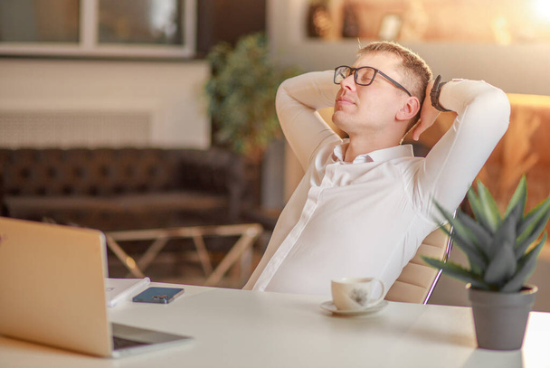Een zakenman rust op zijn werkplek met zijn handen achter zijn hoofd, een man met een bril, een kantoormedewerker, een afdelingshoofd, een pauze in het werk, een pauze en rust, ontspanning. - Foto, afbeelding