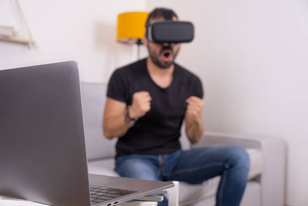 Jeune homme en VR lunettes casque à la maison avec ordinateur portable, future technologie, éducation jeux vidéo - Photo, image