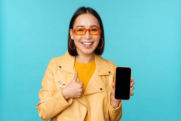 Joven mujer china elegante que muestra la pantalla del teléfono móvil y pulgares hacia arriba, aplicación para teléfonos inteligentes, de pie sobre fondo azul - Foto, imagen
