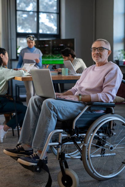 Rijpe werknemer met laptop op zijn knieën zittend in rolstoel - Foto, afbeelding