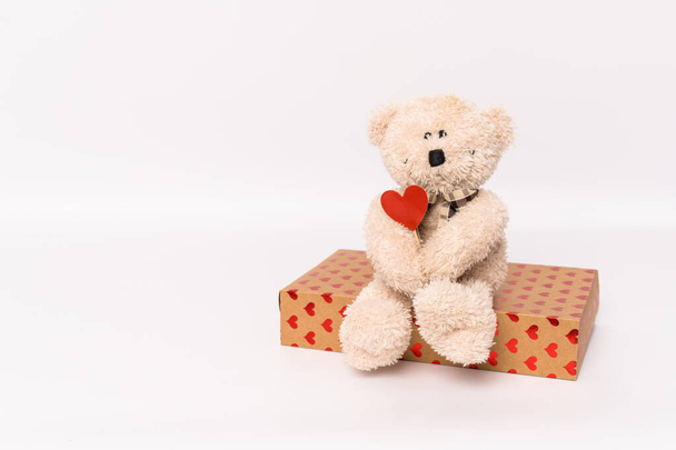 Teddy bear is holding a red heart sitting on gift box isolated over white background. Love concept. - Fotoğraf, Görsel