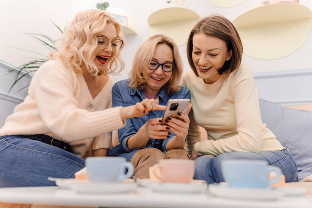 Three woman on a coffee break watching news on a phone. Coleagues at work eat lunch and drink tea together - Foto, imagen