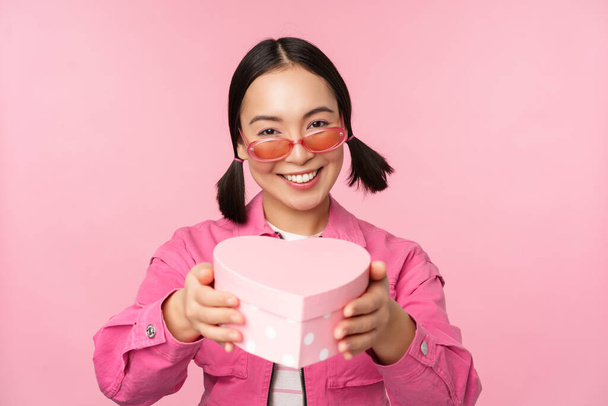 Χαριτωμένο κορίτσι της Ασίας που σας δίνει δώρο σε σχήμα καρδιάς κουτί, φιλιά και χαμόγελα, έννοια των διακοπών και των εορτασμών, στέκεται πάνω από ροζ φόντο - Φωτογραφία, εικόνα