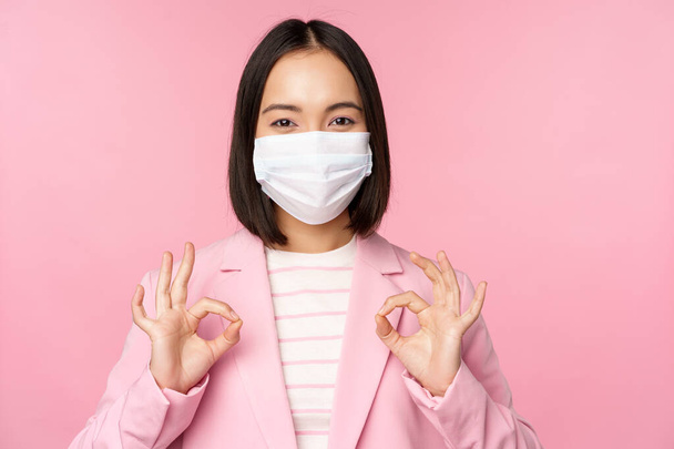 Asian businesswoman in suit and medical face mask, showing okay sign, recommending using protective equipment in office during covid-19 pandemic, pink background - Фото, изображение