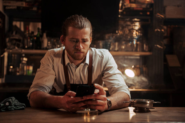 Fiatal kaukázusi férfi csapos egyedül ül a bárban és mobileszközön SMS-ezik.  - Fotó, kép