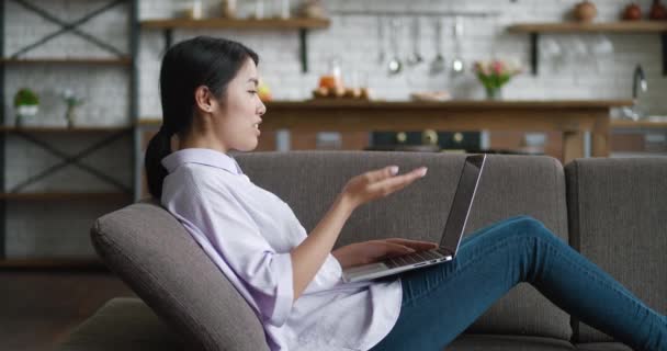 Asian business woman video chatting with laptop on couch at home. Female student talking and looking at laptop screen while resting on couch at home living room. Webcam webinar concept - Footage, Video