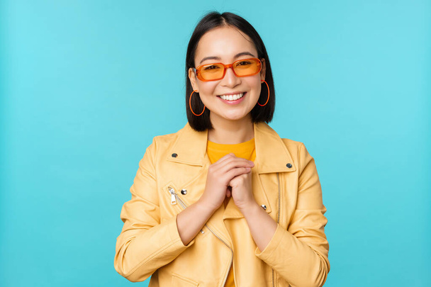 Güneş gözlüklü Asyalı bir kadının portresi, umut dolu, pohpohlanmış, mutlu, mavi arka planda duran - Fotoğraf, Görsel