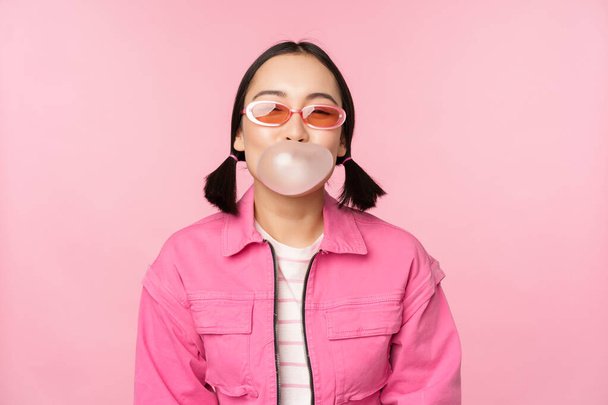 スタイリッシュなアジアの女の子吹いてバブルガムバブル,ガムを噛む,サングラスをかけて,ピンクの背景に対してポーズ - 写真・画像