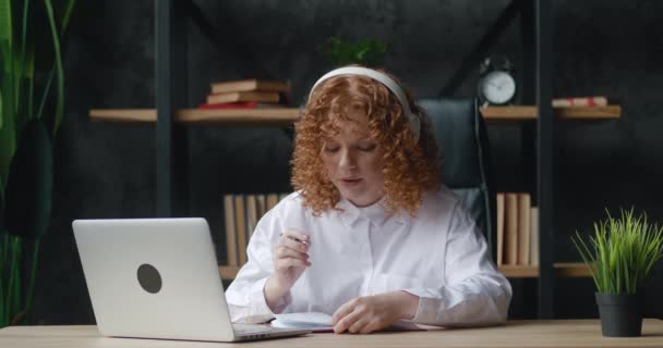 Молода студентка червоно-волохата жінка з навчанням гарнітурі з онлайн-вчителькою розмовляє відеоконференцією, виклик відеочату робить нотатки сидіти на домашньому офісному столі дивіться на екрані комп'ютера ноутбука
 - Кадри, відео