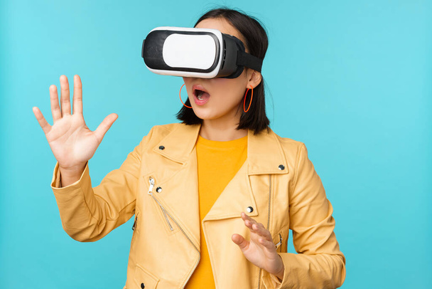 Młoda Azjatka w okularach wirtualnej rzeczywistości, korzystająca z zestawu słuchawkowego VR, bawiąca się na niebieskim tle - Zdjęcie, obraz