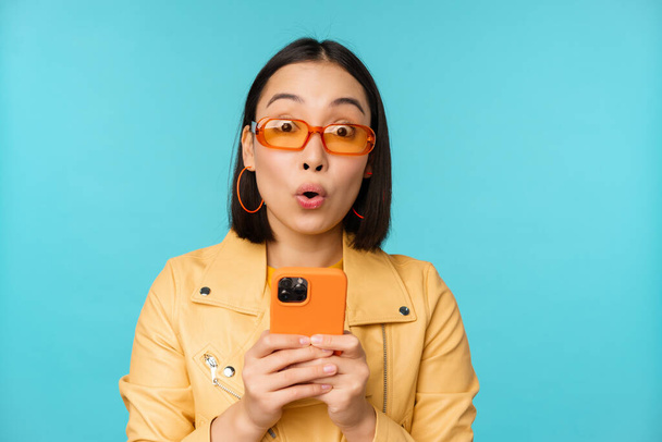 Image of asian girl in sunglasses, looking amazed and impressed, recording video or taking photo on smartphone, standing over blue background - Foto, Imagen
