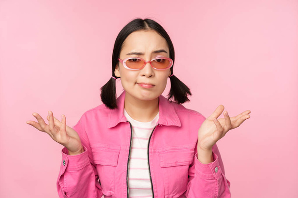 Крупный план портрета азиатской девушки, выглядящей смущенной, пожимающей плечами и смотрящей в камеру, в солнечных очках, стоящей на розовом фоне - Фото, изображение