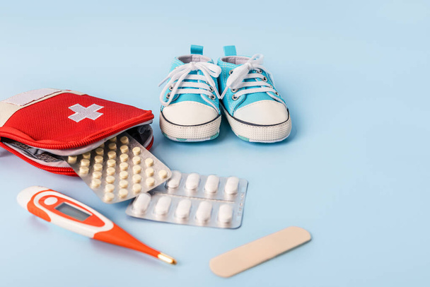 Children's shoes and a first aid kit with a thermometer and pills on a blue background. Space for text. The concept of children's health, treatment - Photo, Image