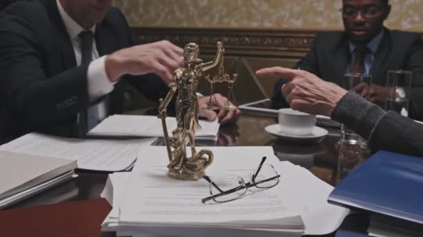 Zbliżenie zdjęcia posągu Themis i okularów na stosie dokumentów przy stole konferencyjnym. Prawnicy prowadzący dyskusję w tle - Materiał filmowy, wideo