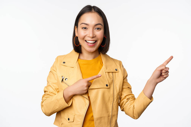 Захоплена азіатська дівчина спрямовує пальці праворуч, показуючи рекламу і посміхаючись, запрошуючи в магазин, стоячи на білому тлі
 - Фото, зображення