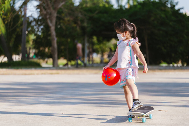 Portrait of Kid girl playing skateboard on street. Child holding red ball. Children wearing white face mask. Happy time. New Normal. Baby aged 5 years old. - Fotó, kép
