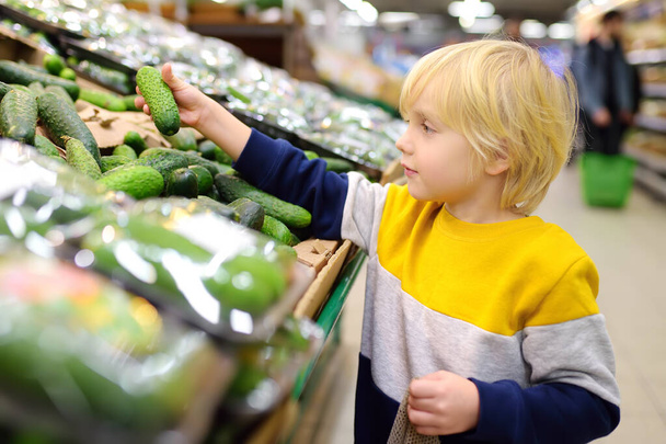 Cute little boy in a food store or a supermarket choosing fresh organic cucumbers. Healthy vegetables for family with kids. Shopping with child. - Photo, Image