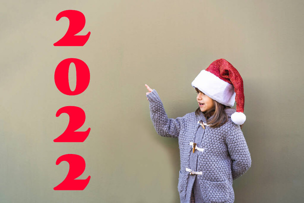 Mutlu küçük kız Noel Baba şapkası takıp 2022 yılının yeşil arka planında duran fotokopi alanını işaret ediyor. - Fotoğraf, Görsel