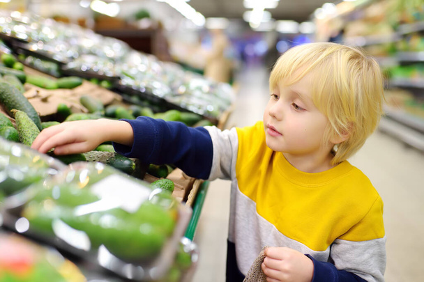 Cute little boy in a food store or a supermarket choosing fresh organic cucumbers. Healthy vegetables for family with kids. Shopping with child. - Photo, Image