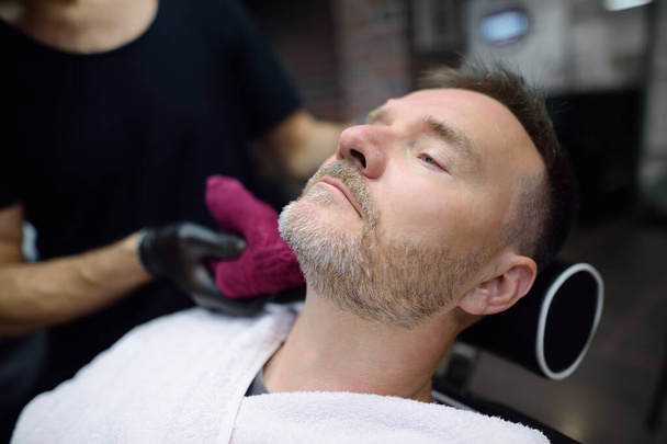 Barber master shaving handsome mature bearded man in salon. Hair artist making beard style for person in male barbershop. Services of professional stylist. Fashion haircare for men - Photo, image