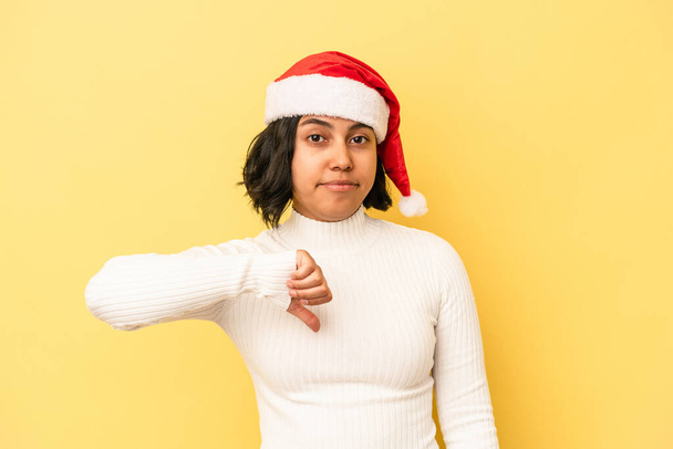黄色の背景に隔離されたクリスマスを祝う若いラテン語の女性は嫌なジェスチャー、親指を下に示す。意見の相違. - 写真・画像
