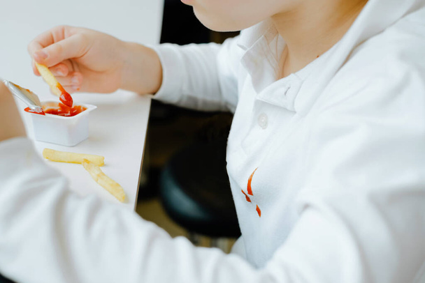 Fechar as manchas sujas de ketchup em roupas de criança branca. Menino comendo batatas fritas batatas fritas e molho de tomate na mesa em um restaurante de fast food. Crianças comem alimentos gordurosos não saudáveis.  - Foto, Imagem