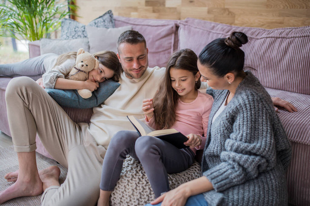 Two happy sisters with mother and father sitting on floor hugging and reading book at home - Фото, зображення