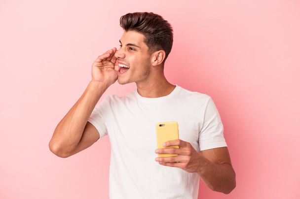 Jovem caucasiano segurando um telefone celular isolado em fundo rosa gritando e segurando palma perto da boca aberta. - Foto, Imagem