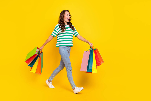 Full size profile photo of impressed brunette millennial lady go with bags wear t-shirt jeans sneakers isolated on yellow background - Foto, Imagen