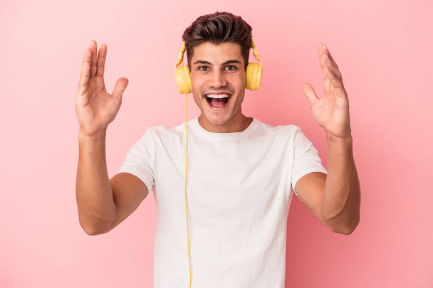 Young caucasian man listening to music isolated on pink background receiving a pleasant surprise, excited and raising hands. - Fotó, kép