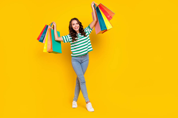 Full body photo of cool brunette young lady hold bags wear t-shirt jeans sneakers isolated on yellow background - Foto, Imagem