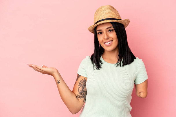 Jonge blanke vrouw met een arm met een zomerhoed op een roze achtergrond die een kopieerruimte op een palm toont en een andere hand op de taille houdt. - Foto, afbeelding